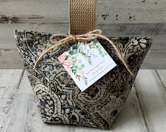 Peut inclure: Un cale-porte en tissu avec un motif damassé noir, gris et beige et une poignée tissée beige. Une étiquette carrée blanche attachée avec de la ficelle présente une illustration florale et le texte DS Design and Textiles, handcrafted designer pillows, etc.