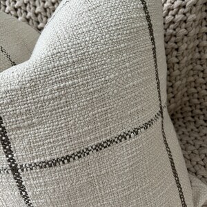 May include: A close-up of a cream-colored throw pillow with a subtle black and white checkered pattern. The pillow is made of a textured fabric and has a soft, cozy feel.