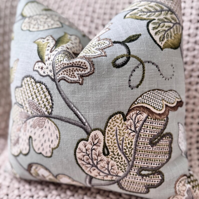 Peut inclure: Un coussin bleu clair avec un motif floral dans les tons de vert, marron et blanc. Le coussin pr&eacute;sente un d&eacute;licat motif brod&eacute; de feuilles et de vignes.