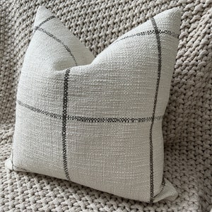 May include: A cream-colored throw pillow with a subtle black and white checkered pattern. The pillow is made of a soft, textured fabric.