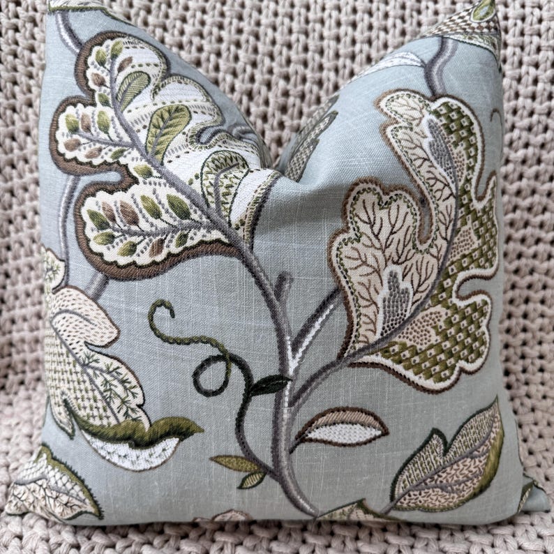 Peut inclure: Un coussin bleu clair avec un motif floral brod&eacute; dans des tons de vert, marron et beige. Le coussin pr&eacute;sente un motif feuillu avec des d&eacute;tails complexes.