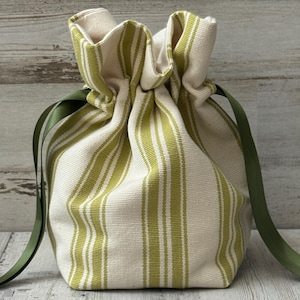 May include: A drawstring bag made of cream-colored fabric with vertical green stripes. The bag is gathered at the top and tied with a green ribbon. The bag is likely used for storage or as a gift bag.