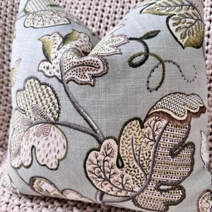Peut inclure: Un coussin bleu clair avec un motif floral dans les tons de vert, marron et blanc. Le coussin pr&eacute;sente un motif r&eacute;p&eacute;titif de feuilles et de vignes stylis&eacute;es.