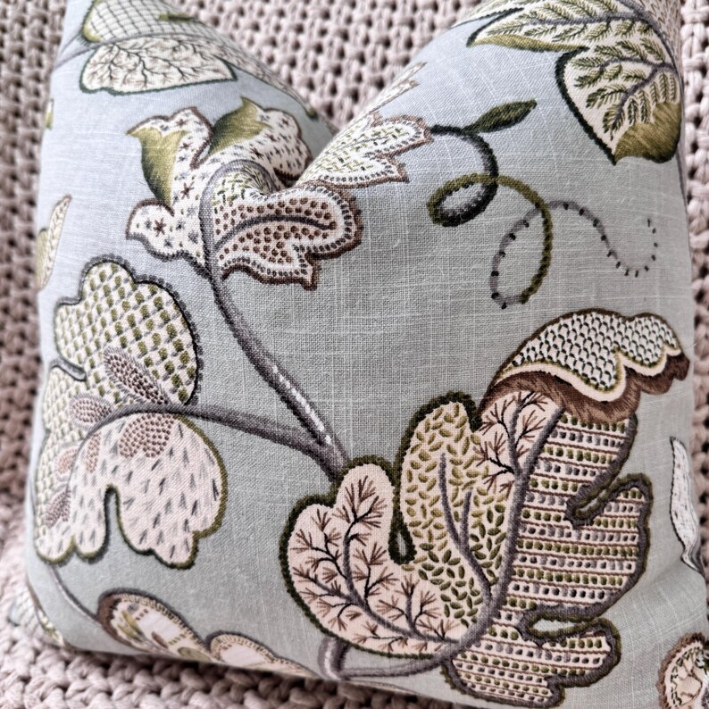 Peut inclure: Un coussin bleu clair avec un motif floral dans les tons de vert, marron et blanc. Le coussin pr&eacute;sente un motif r&eacute;p&eacute;titif de feuilles et de vignes stylis&eacute;es.