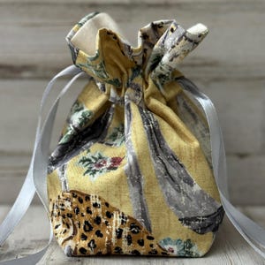 May include: A small, drawstring gift bag with a yellow background and a mix of patterns. The patterns include a leopard print, floral designs, and vertical gray stripes. The bag is tied with a silver ribbon.