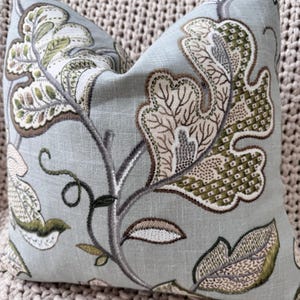 Peut inclure: Un coussin bleu clair avec un motif floral dans les tons de vert, marron et beige. Le coussin pr&eacute;sente un motif r&eacute;p&eacute;titif de feuilles et de branches stylis&eacute;es.