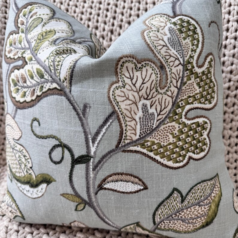 Peut inclure: Un coussin bleu clair avec un motif floral dans les tons de vert, marron et beige. Le coussin pr&eacute;sente un motif r&eacute;p&eacute;titif de feuilles et de branches stylis&eacute;es.