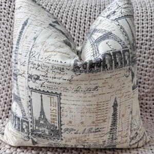 May include: A white and black pillow with a Parisian theme. The pillow features a repeating pattern of the Eiffel Tower, handwritten script, and postage stamps. The pillow is made of a soft, textured fabric.
