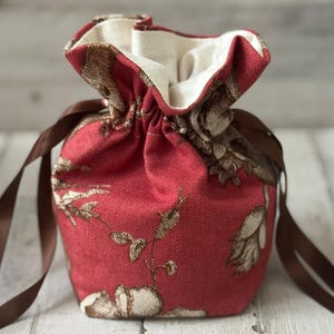 May include: A red fabric drawstring bag with a floral pattern in brown and cream. The bag has a cream-colored interior and brown ribbon ties. The bag is designed to be closed by pulling the ribbon.