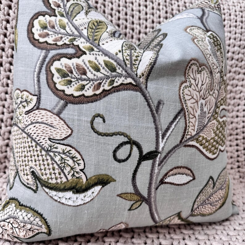 Peut inclure: Un coussin bleu clair avec un motif floral brod&eacute; dans des tons de vert, marron et rose. Le coussin pr&eacute;sente un motif r&eacute;p&eacute;titif de feuilles et de fleurs.