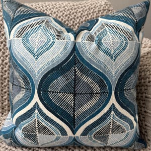 May include: Square decorative pillow with a geometric pattern in shades of blue, white, and black. The design features a repeating motif of stylized leaf shapes, creating a symmetrical and visually appealing pattern. The pillow is placed on a textured, neutral-colored surface.