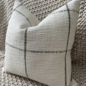 May include: A cream-colored throw pillow with a subtle brown plaid pattern. The pillow is made of a textured fabric and has a square shape.