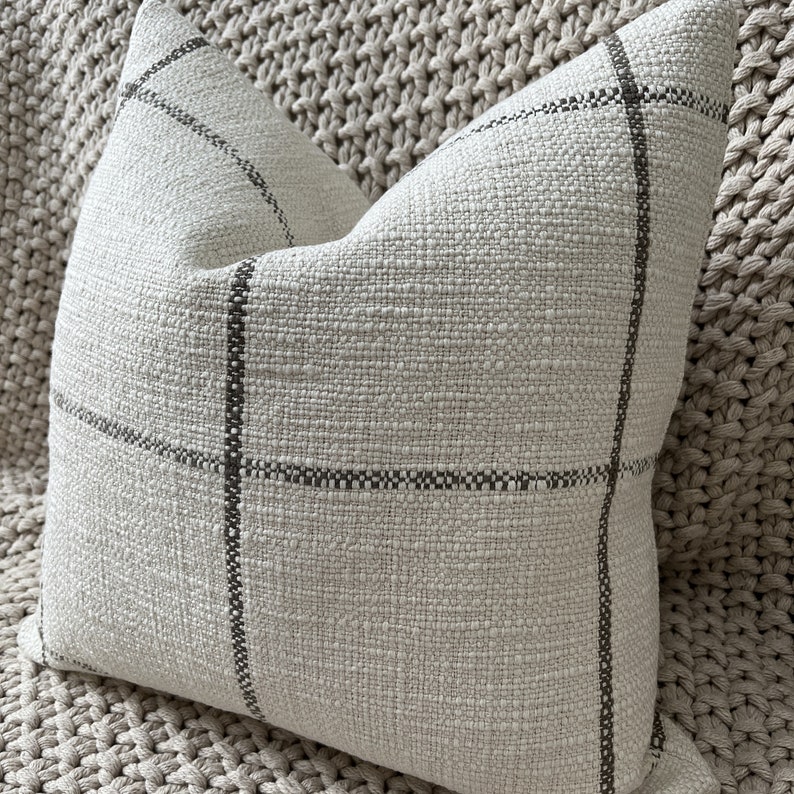 May include: A cream-colored throw pillow with a subtle brown plaid pattern. The pillow is made of a textured fabric and has a square shape.