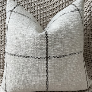 May include: A white throw pillow with a gray plaid pattern. The pillow is made of a soft, textured fabric.