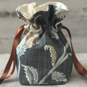 May include: A small, drawstring bag with a floral pattern in shades of gray, brown, and cream. The bag is tied with a brown ribbon. The interior lining is a cream color.