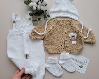 May include: A knitted baby outfit set in white and brown. The set includes a white knitted hat, a brown knitted cardigan with a bear applique, white knitted overalls, and white knitted socks.