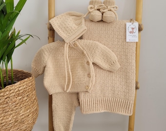 May include: A beige knitted baby outfit set with a hat, cardigan, pants, and booties. The set is made of soft, textured yarn and has a subtle dot pattern.