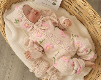 May include: A knitted baby outfit in a light pink color with a floral design. The outfit includes a hat, a long-sleeved romper, and booties. The romper has a ruffle at the shoulders and a bow at the neck. The outfit is decorated with pink flowers and green leaves.
