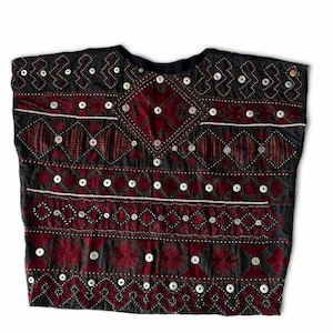 Peut inclure: Un haut noir sans manches orné de bandes horizontales de broderies rouge foncé aux motifs géométriques en losanges et en zigzag. Le vêtement est agrémenté de nombreux petits boutons ronds blancs disposés en rangées sur le devant.