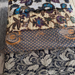 May include: A stack of five folded fabric pieces featuring intricate batik patterns. The designs include floral motifs, geometric shapes, and paisley elements in a colour palette of cream, brown, navy blue, black, and accents of purple and bright blue.