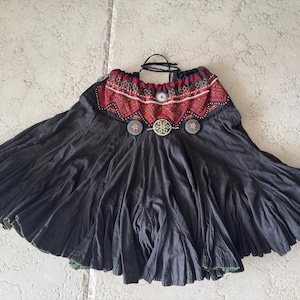 Puede incluir: Una falda negra acampanada con una cintura ancha y fruncida que presenta un patrón geométrico en rojo y negro. La cintura está decorada con tres botones circulares de tono metálico. La falda tiene una silueta amplia y plisada.