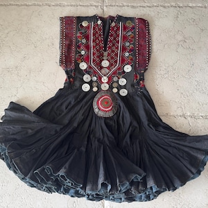 Peut inclure: Une robe noire sans manches avec une jupe évasée et plissée ainsi qu'un corsage décoratif. La partie supérieure est ornée de broderies rouges complexes, de surpiqûres blanches et de nombreux disques et boutons métalliques argentés.