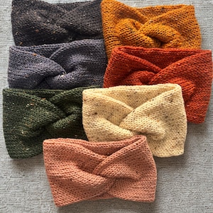 May include: Seven knitted headbands in a variety of colours, including dark grey, mustard yellow, rust orange, olive green, cream, and peach. Each headband has a twisted knot design. The headbands are made of textured yarn.