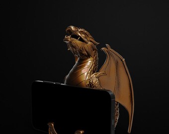 May include: A bronze-coloured dragon-shaped phone holder. The dragon has detailed scales, wings, and an open mouth. The phone rests in a slot at the base, with the dragon's body supporting the phone. The background is black.