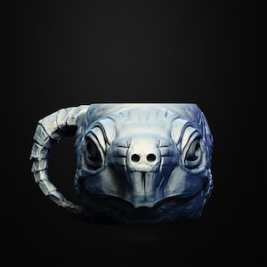 May include: A ceramic mug shaped like a turtle's head, with a textured, gray-blue finish. The handle is designed to resemble the turtle's neck. The mug has detailed facial features, including eyes and nostrils, and is set against a black background.