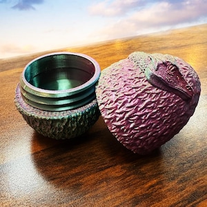 Puede incluir: Un recipiente de dos partes con diseño de reptil. La tapa es de forma esférica texturizada en tonos morados y rosas, con una cabeza de reptil esculpida en la parte superior. La base es cilíndrica con un exterior estriado.