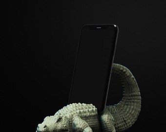May include: A light green alligator phone holder with a black smartphone. The alligator is sculpted with textured scales and a curved tail. The phone is held upright, resting against the alligator's back.
