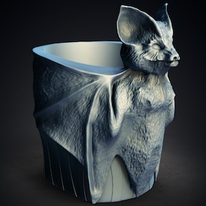 May include: A gray ceramic cup shaped like a bat with its wings folded around the cup. The bat has a detailed face and fur texture.