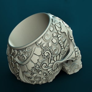May include: A white ceramic skull planter featuring intricate floral designs. The planter has a wide opening at the top and is designed to hold small plants or succulents.