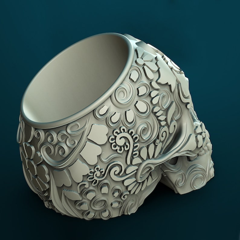 May include: A white ceramic skull planter featuring intricate floral designs. The planter has a wide opening at the top and is designed to hold small plants or succulents.