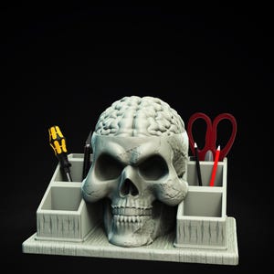 May include: A gray skull-shaped desk organizer with a brain-like interior. It features compartments holding a yellow and black screwdriver, a black pen, a red pencil, and red scissors. The base and compartments have a textured, weathered appearance.