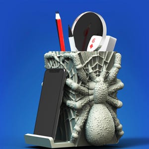 Könnte beinhalten: Ein grauer Spinnen-Schreibtisch-Organizer mit einem Spinnennetz-Design. Er hat einen Telefonhalter, einen Stifthalter und ein kleines Fach für andere Gegenstände.