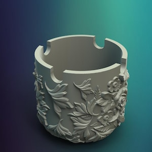 May include: A light gray ashtray with a floral and leaf design. The ashtray has three notches for holding cigarettes and is made of a solid material. The floral design is carved into the side of the ashtray.