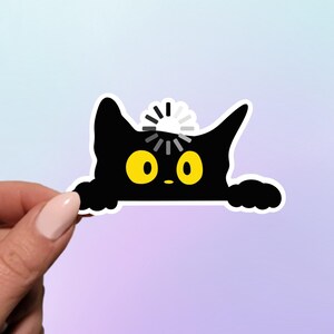 May include: A black cat sticker with yellow eyes and a white loading symbol above its head. The cat is peeking over a surface. The sticker is held by a hand with a light pink nail polish.