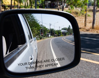 May include: A car's side mirror reflecting a winding road with yellow and white lane markings. The text "YOUR DEMONS ARE CLOSER THAN THEY APPEAR" is printed in white on the mirror's surface. The sky is clear and blue.