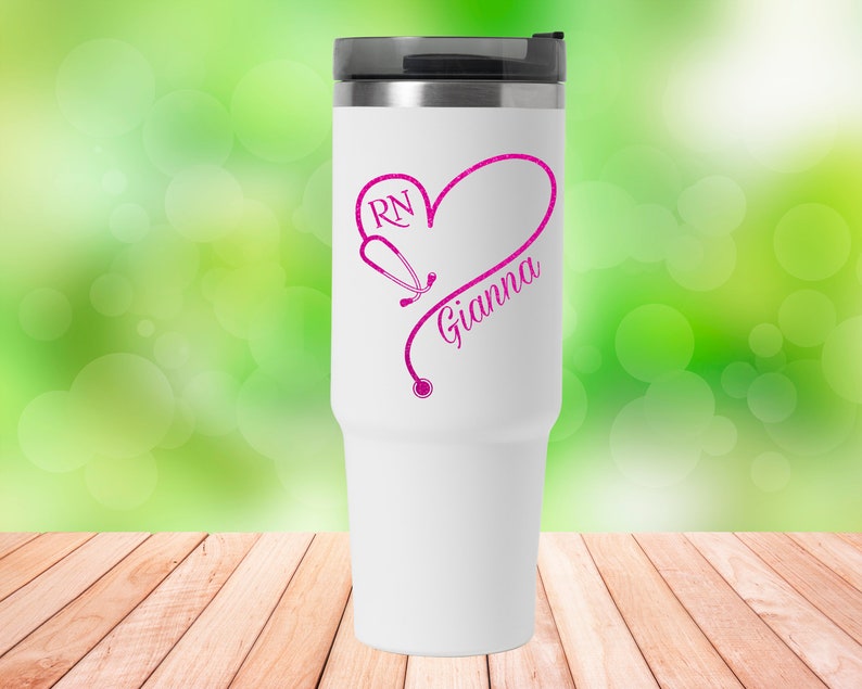 May include: White travel mug with a pink heart design featuring a stethoscope and the text "RN Gianna".
