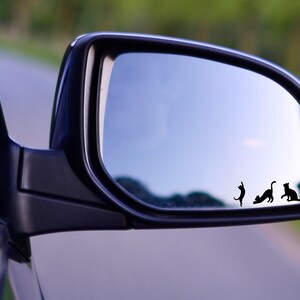 Puede incluir: Un espejo retrovisor con tres siluetas de gatos negros reflejadas en el vidrio. Los gatos est&aacute;n en diferentes poses, uno est&aacute; saltando, otro est&aacute; sentado y otro est&aacute; caminando.