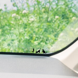 Puede incluir: Una ventana de coche con vista a un campo verde. Hay tres siluetas de gatos negros en la ventana, dos de pie y una sentada.