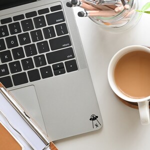 May include: A silver laptop computer with a black keyboard and a black and white sticker of a UFO with an alien inside. The laptop is on a white surface next to a cup of coffee and a jar of pencils.
