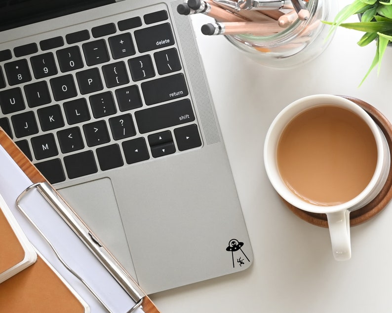 May include: A silver laptop computer with a black keyboard and a black and white sticker of a UFO with an alien inside. The laptop is on a white surface next to a cup of coffee and a jar of pencils.