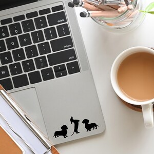 May include: A silver laptop computer with a black keyboard and a black trackpad. Three black dachshund silhouette stickers are on the trackpad. The laptop is on a white surface.