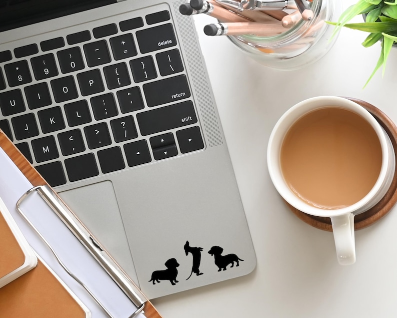 May include: A silver laptop computer with a black keyboard and a black trackpad. Three black dachshund silhouette stickers are on the trackpad. The laptop is on a white surface.