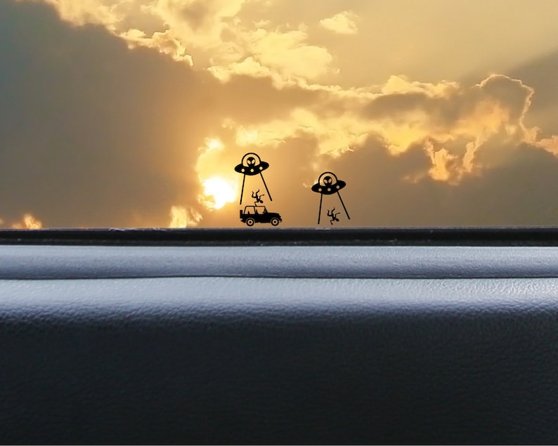 May include: A black car with a silhouette of a UFO hovering above it. The car is driving on a road with a sunset in the background.