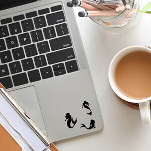 May include: A silver laptop computer with a black keyboard and a black trackpad. Three black mermaid silhouettes are on the trackpad. The laptop is on a white surface with a cup of coffee and a pencil holder.