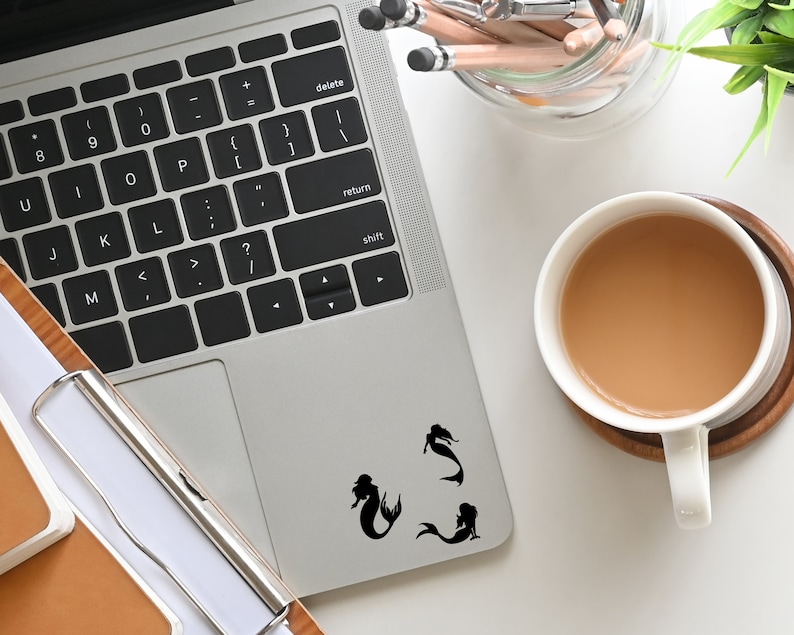 May include: A silver laptop computer with a black keyboard and a black trackpad. Three black mermaid silhouettes are on the trackpad. The laptop is on a white surface with a cup of coffee and a pencil holder.