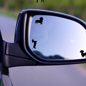 May include: A rearview mirror with three black dachshund silhouette decals. The decals are on the mirror's surface, reflecting the sky and road.
